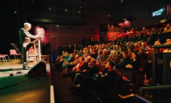 A spot lit man stands on a stage in a grey suit addressing a low lit auditorium full of people
